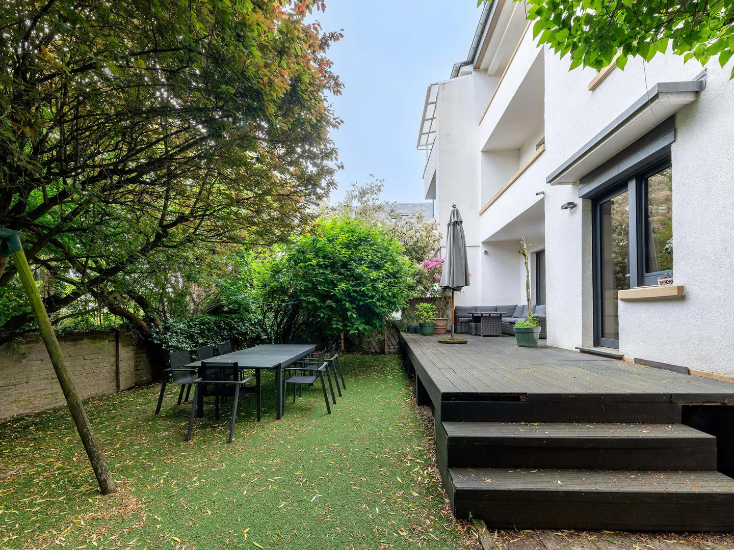 A backyard with a dark wood deck, a table with chairs, and a white house in the background.
