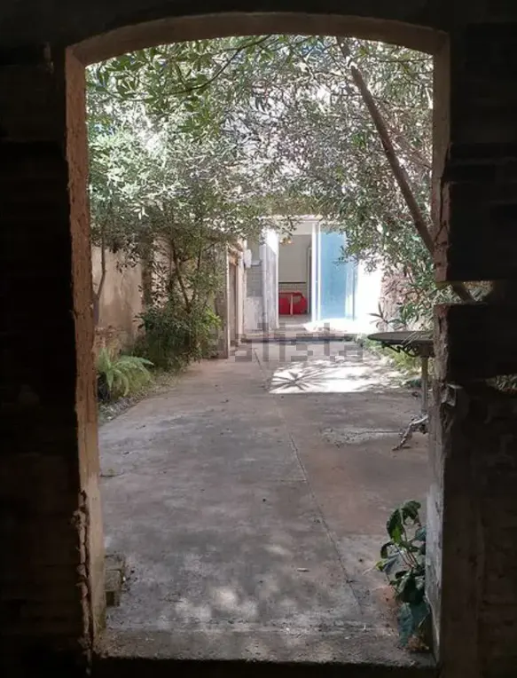 View through a rustic stone archway into a shaded courtyard with trees, a concrete patio, and a bright building entrance.