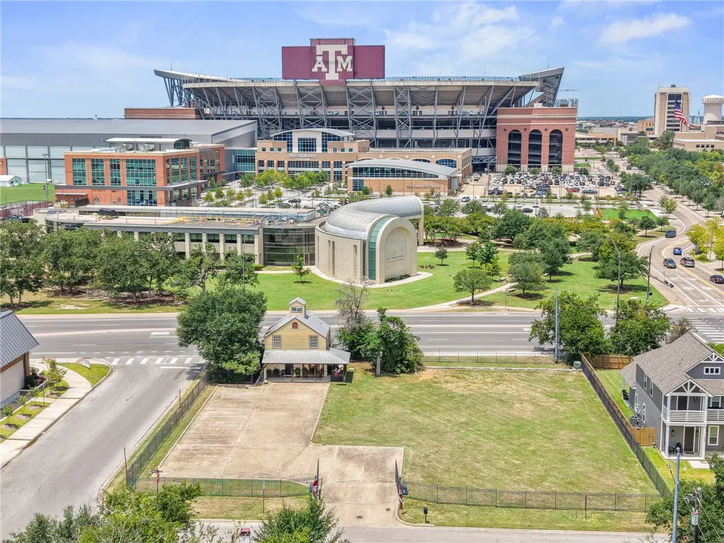 Aerial view of a lot with a yellow house, green grass, and Texas A&M stadium in the background.