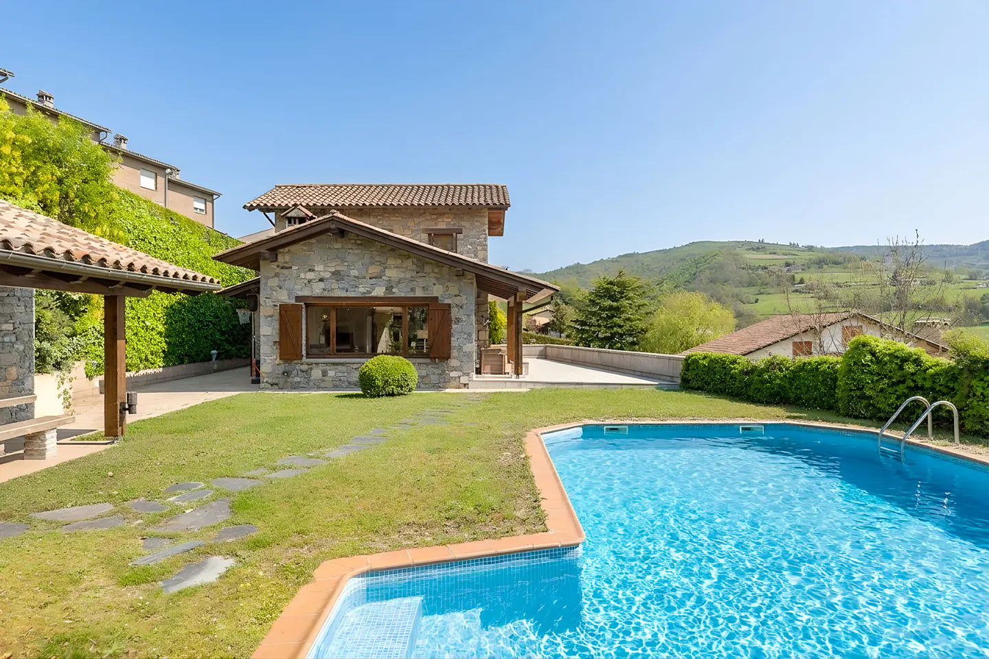 Exterior view of a stone house with a red tile roof, a blue swimming pool, and a green lawn.
