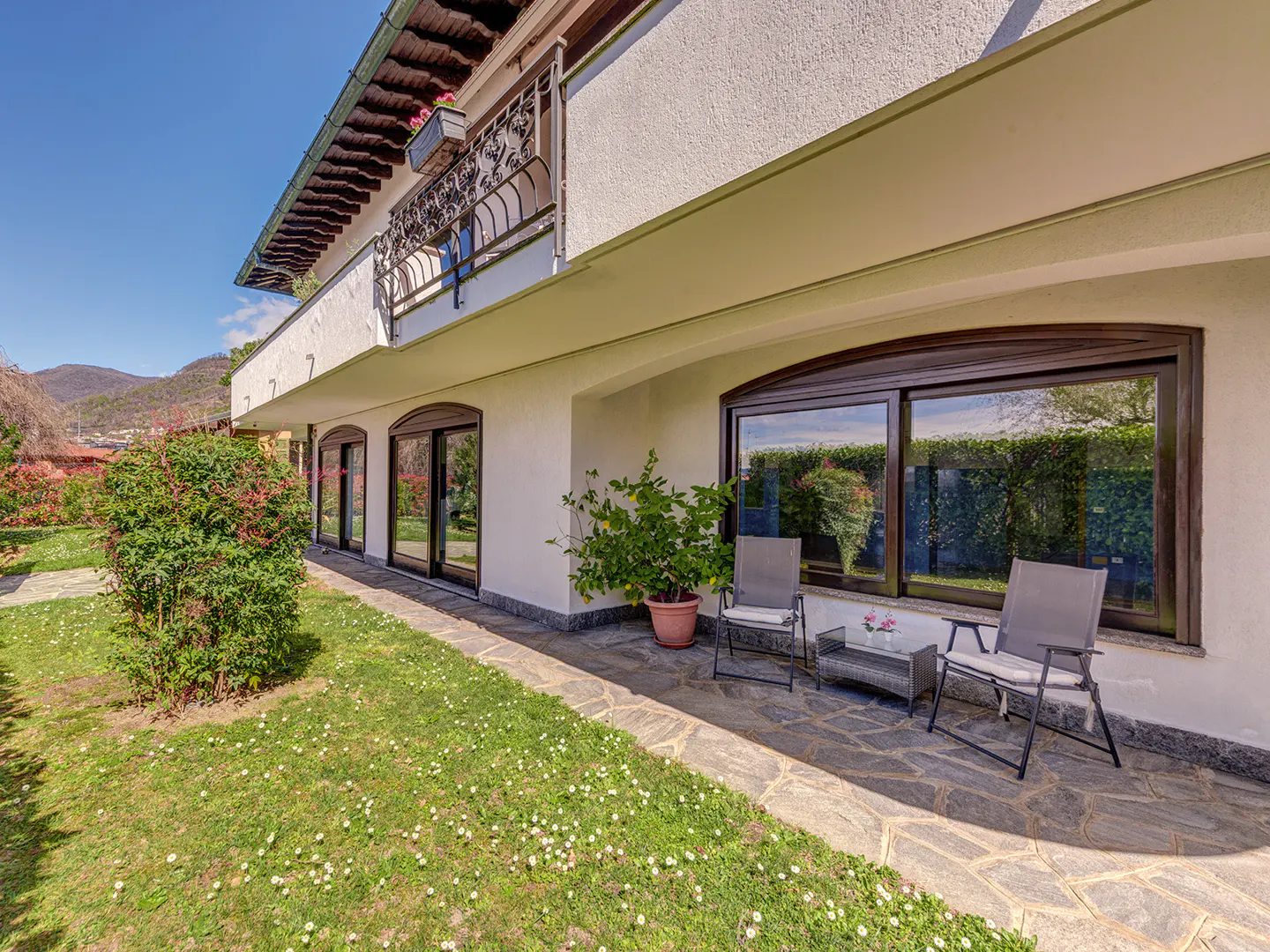 Exterior view of a white house with a balcony, stone patio, and green lawn with white flowers. Two gray chairs and a small table sit on the patio.
