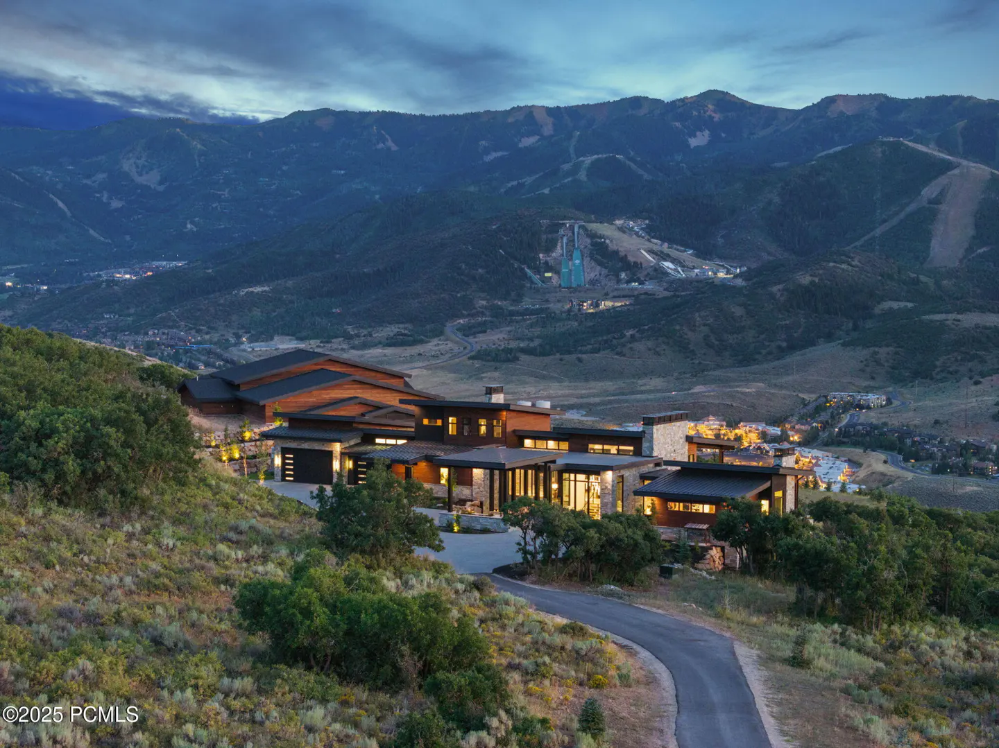 Modern mountain home at dusk with warm interior lighting, nestled on a hillside overlooking a valley and distant mountains.
