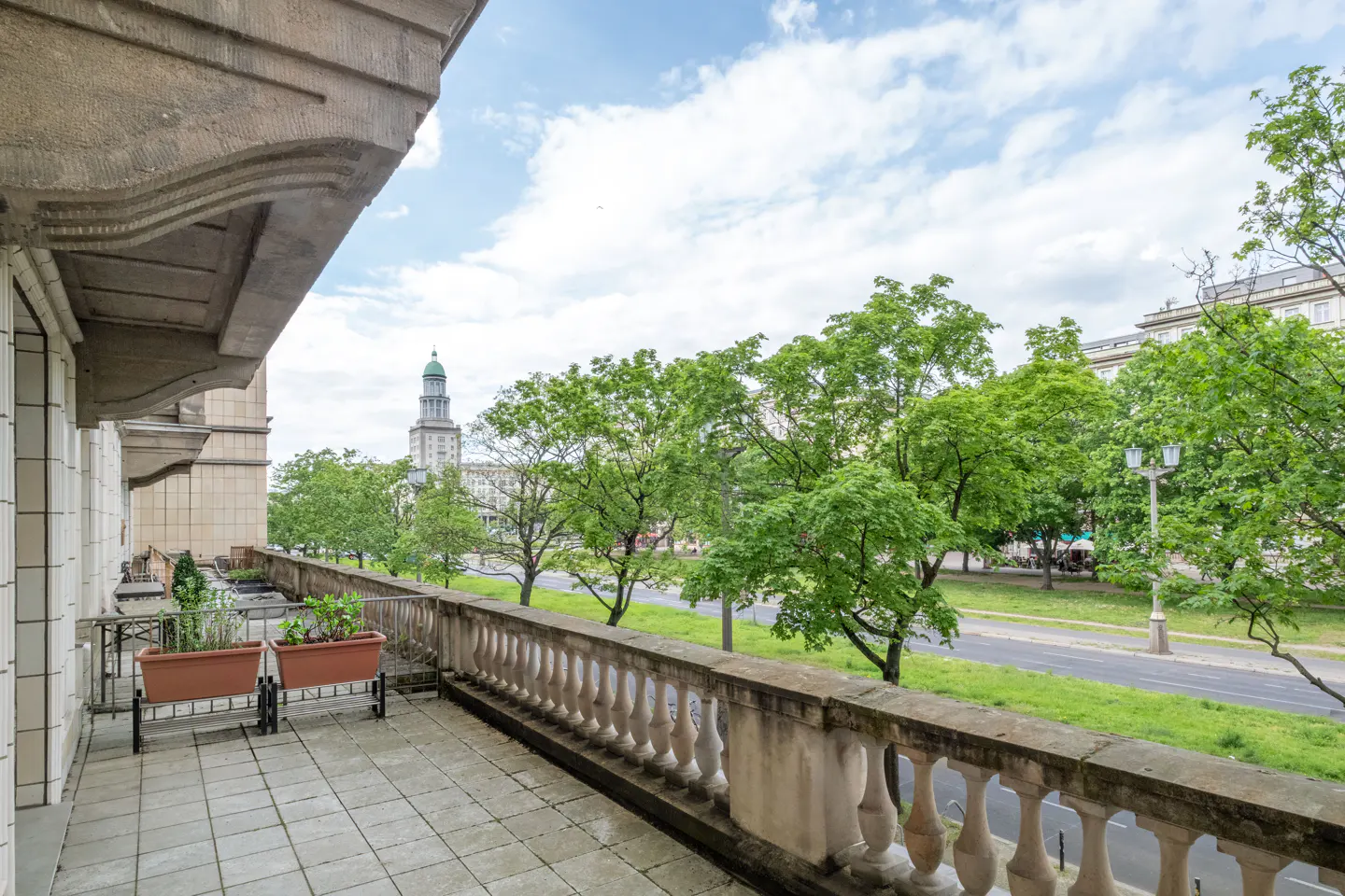 Balcony view with stone railing, potted plants, and a distant building with a green spire, framed by green trees and a cloudy sky.