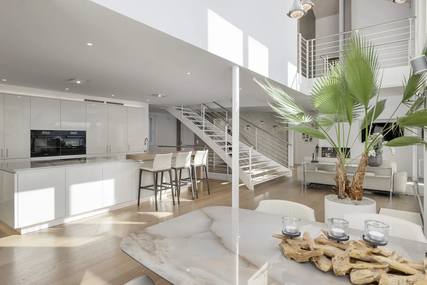 Bright, modern interior with white kitchen, island, and bar stools. A white staircase leads to a second floor living area with a palm tree.