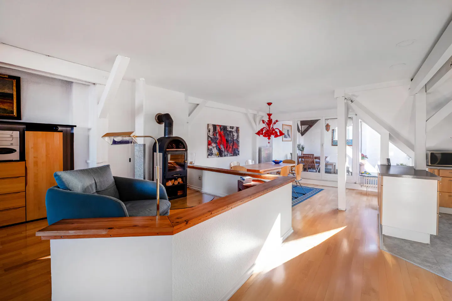 Open-plan living area with a blue armchair, wood-burning stove, dining table, red chandelier, and light wood flooring.