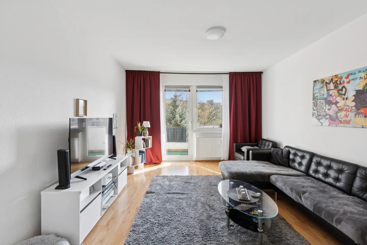 Living room with white walls, wood floors, and a gray rug. A black leather sofa faces a TV on a white console. Red curtains frame a balcony door.
