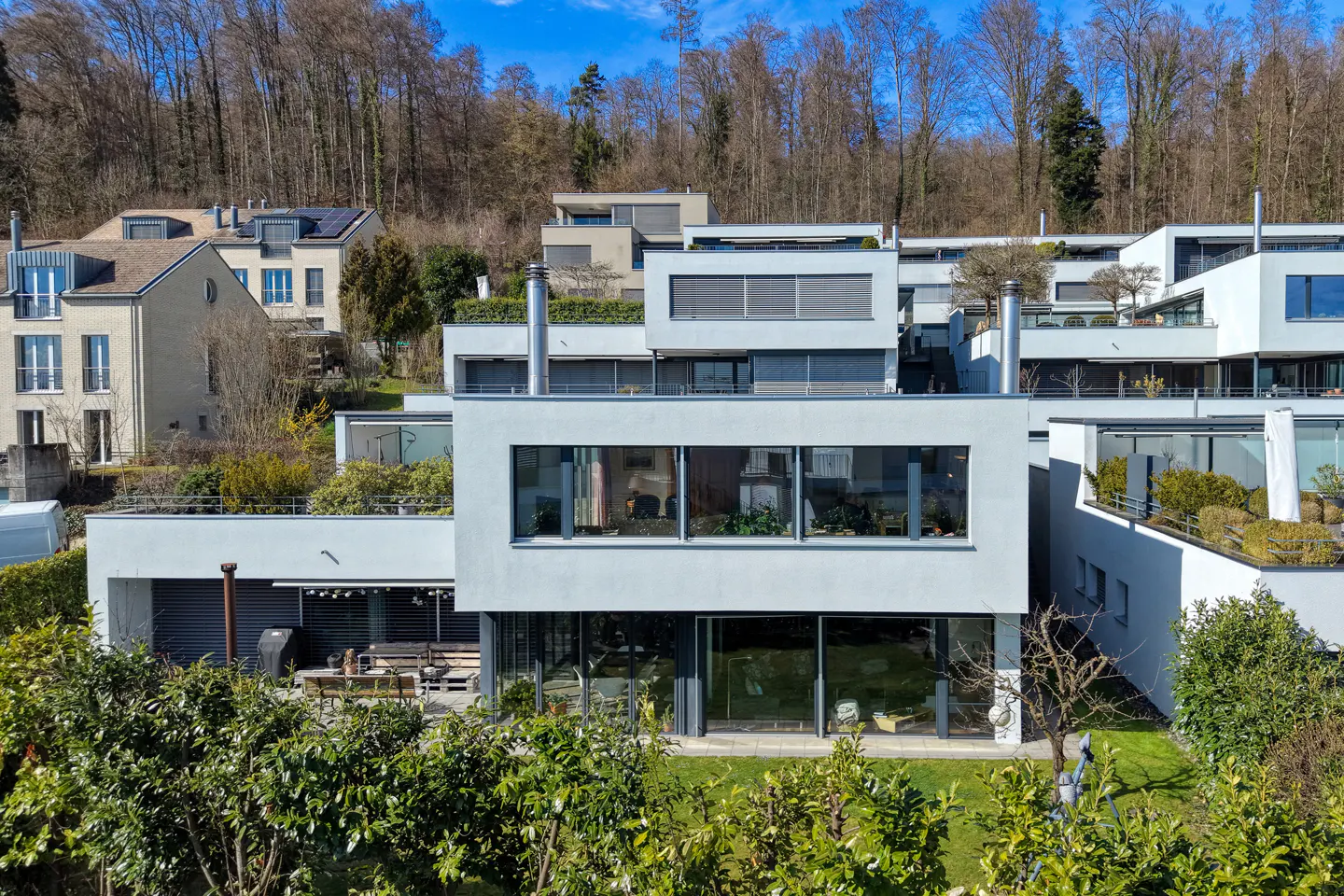 Modern white houses on a hillside, with trees in the background and green bushes in the foreground.