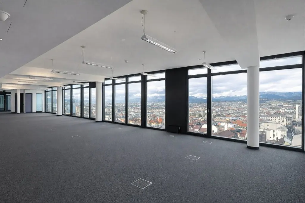 Empty office space with gray carpet, white ceiling, and large windows. Cityscape and mountains visible through the windows.