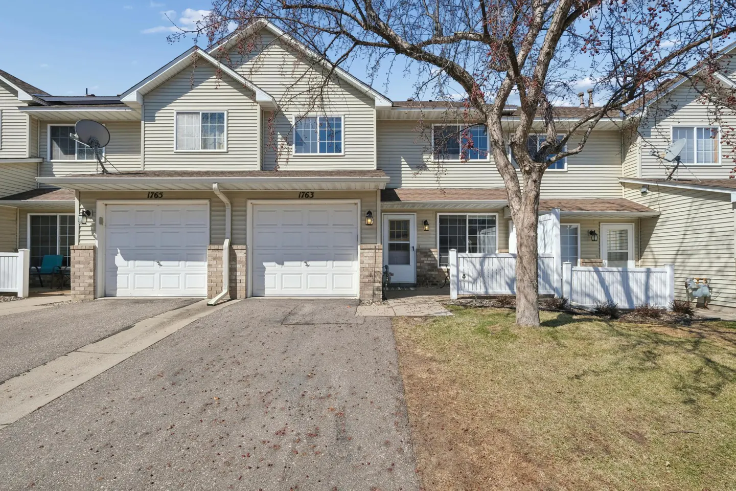 A two-story townhouse with beige siding, white garage doors, and a paved driveway. A tree stands in front of the house.