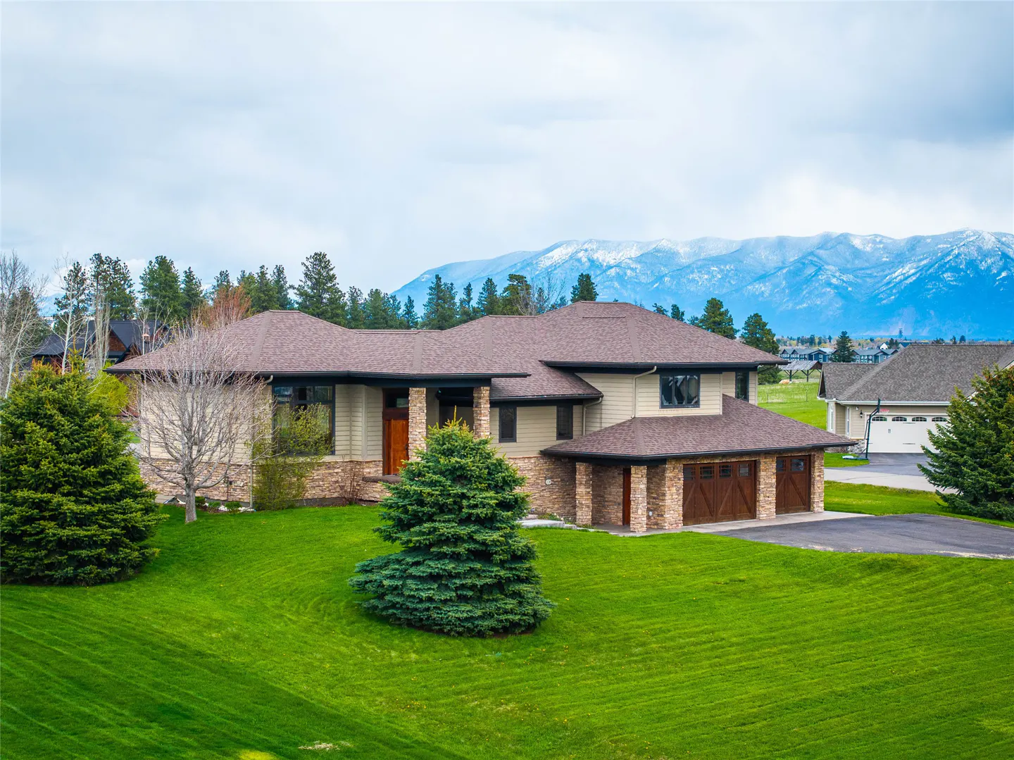 A tan two-story house with a brown roof sits on a green lawn with mountains in the background.