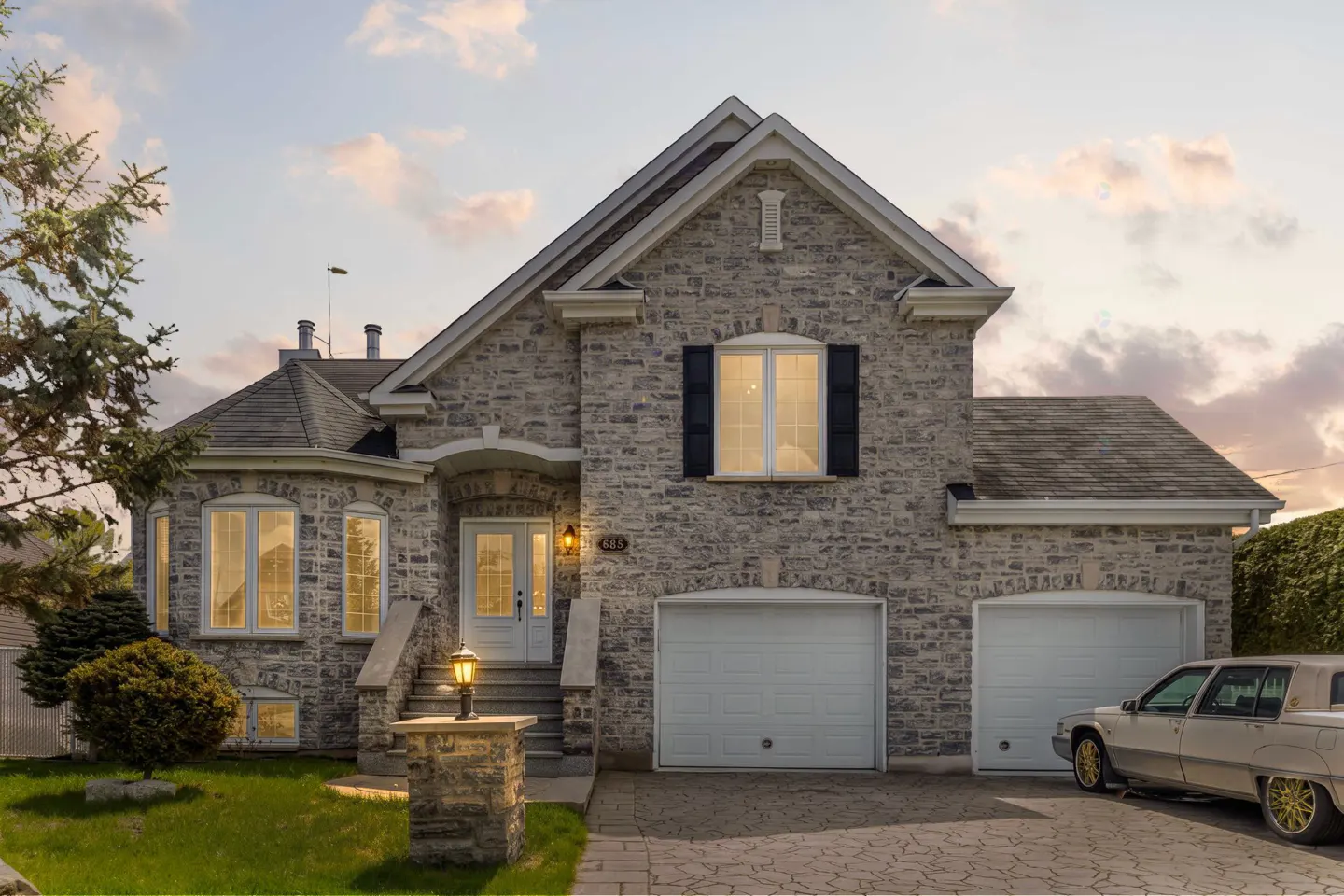 Stone house with two-car garage, lit windows, and a vintage car in the driveway.