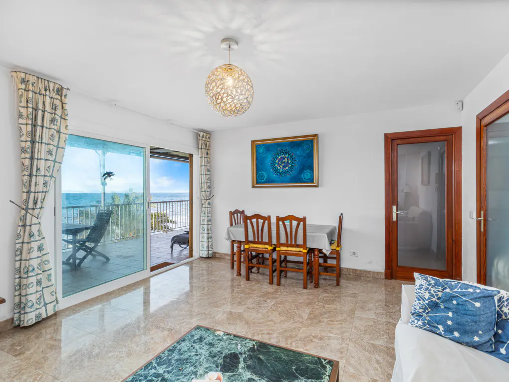 Bright living room with ocean view. Table and chairs sit under a blue painting. Sliding glass doors open to a balcony.