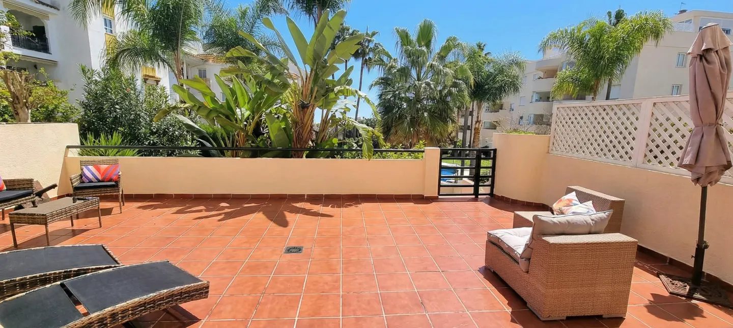 A sunny patio with terracotta tiles, wicker furniture, and lush green plants. A black metal gate leads to a pool. Buildings are visible in the background.