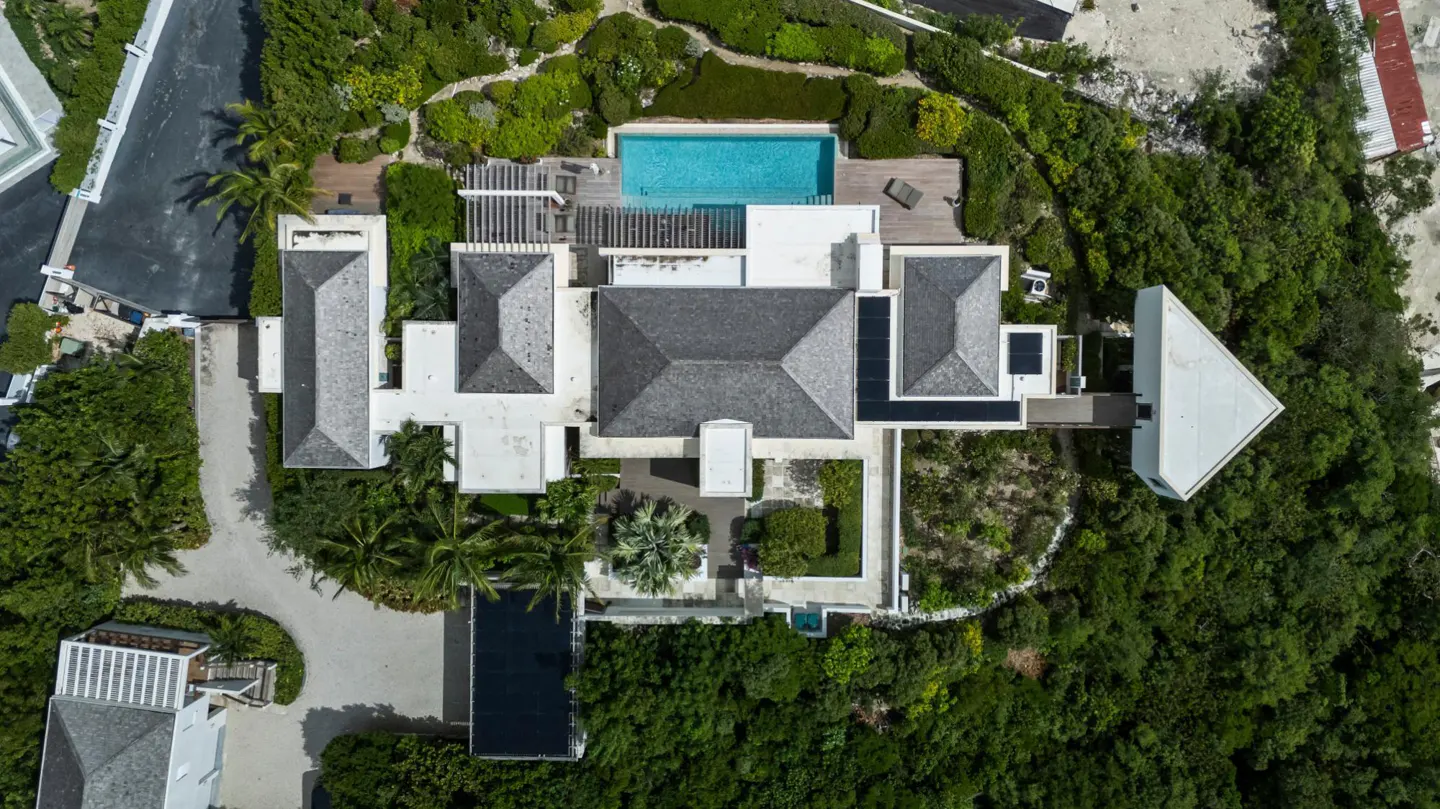Aerial view of a modern white house with a gray roof, surrounded by lush green trees and a blue swimming pool.