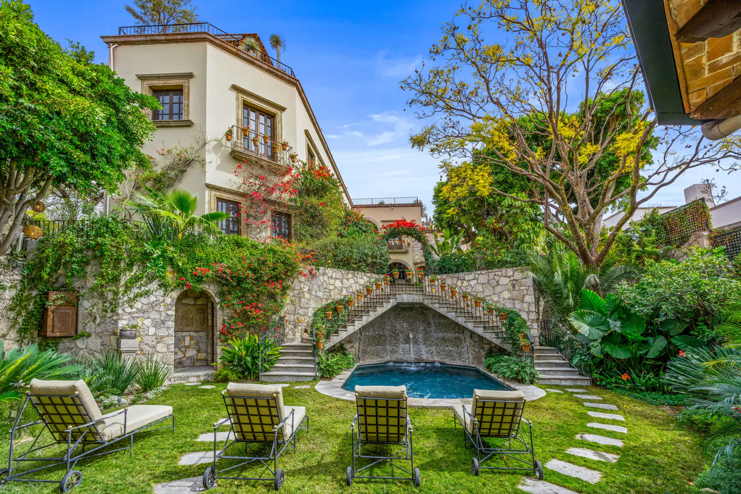 Lounge chairs face a pool with a stone staircase and waterfall feature in a lush garden setting with a cream-colored building backdrop.