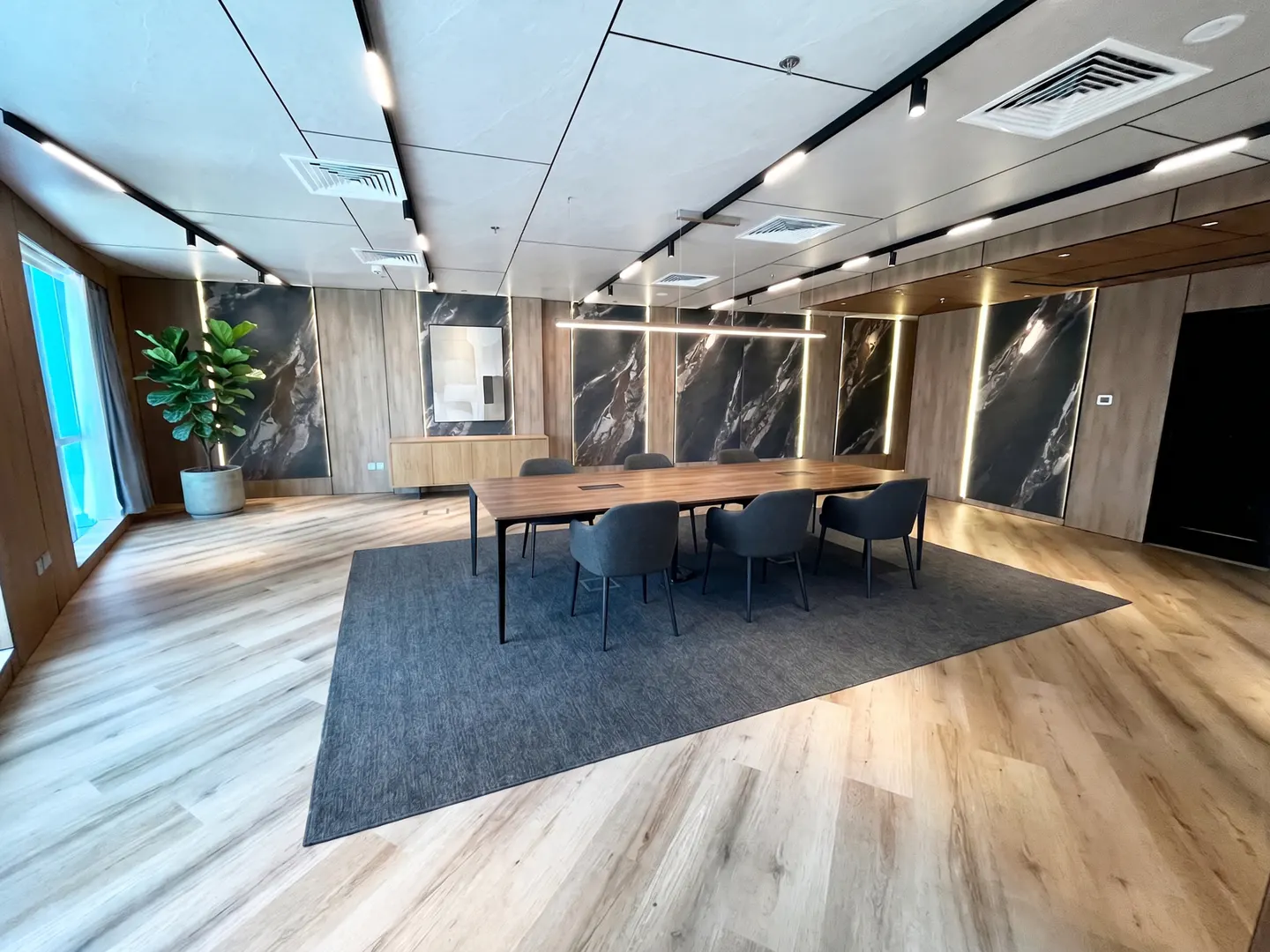 Conference room with a long wooden table, gray chairs, and a gray rug. Marble wall panels with vertical lights. Wood floor and a potted plant by the window.