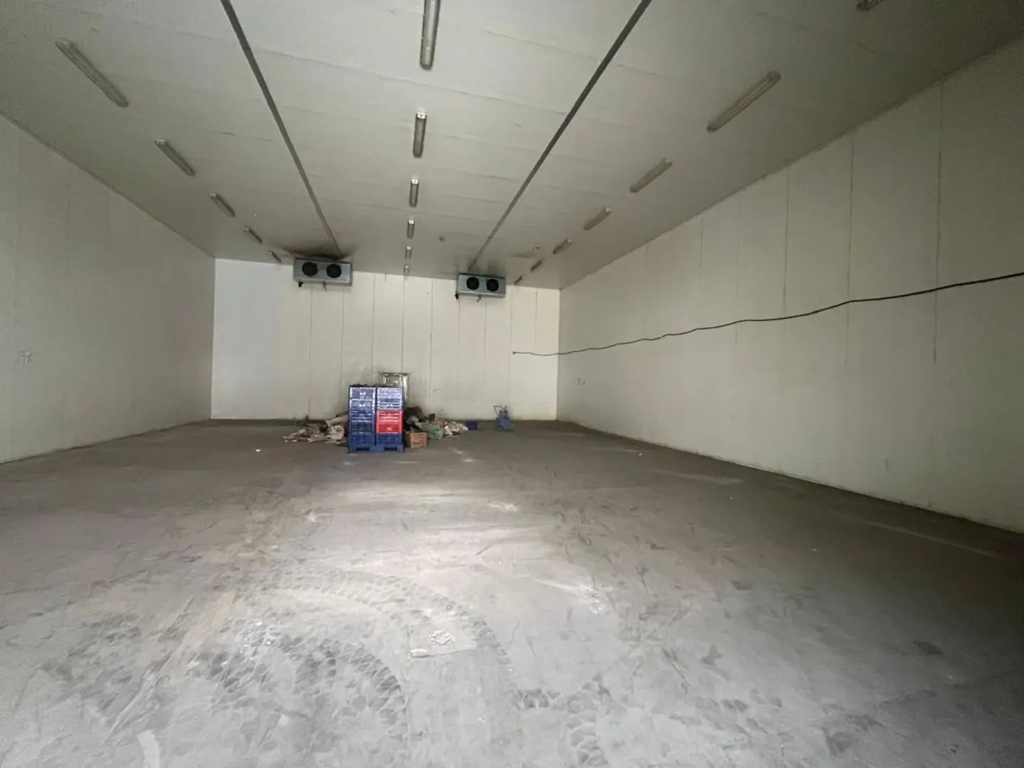 A large, empty warehouse with white walls, a concrete floor with tire tracks, and overhead fluorescent lighting.