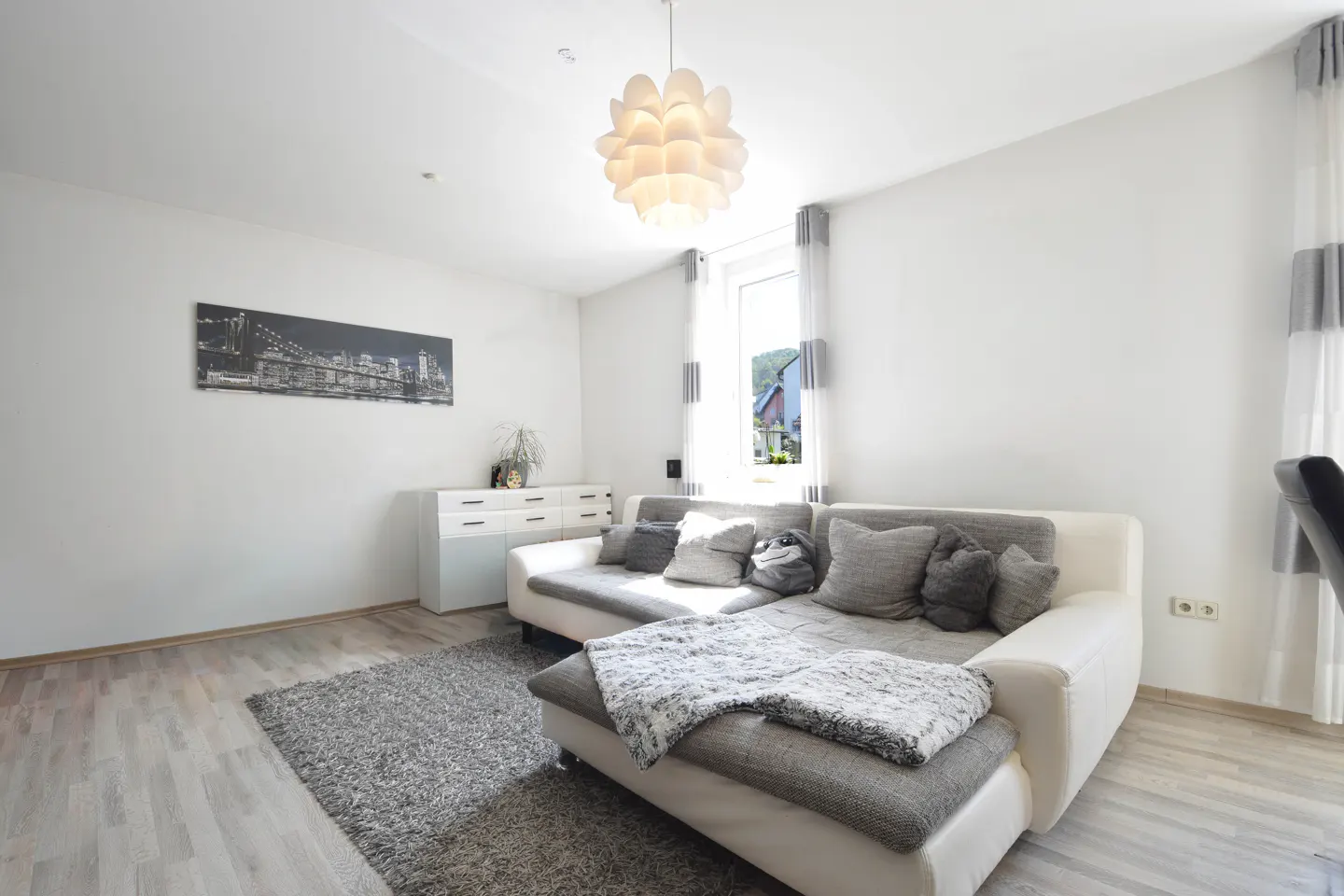 Bright living room with white walls, light wood floors, and a gray rug. A white sectional sofa with gray pillows sits under a modern pendant light. A black and white city print hangs above a white cabinet.