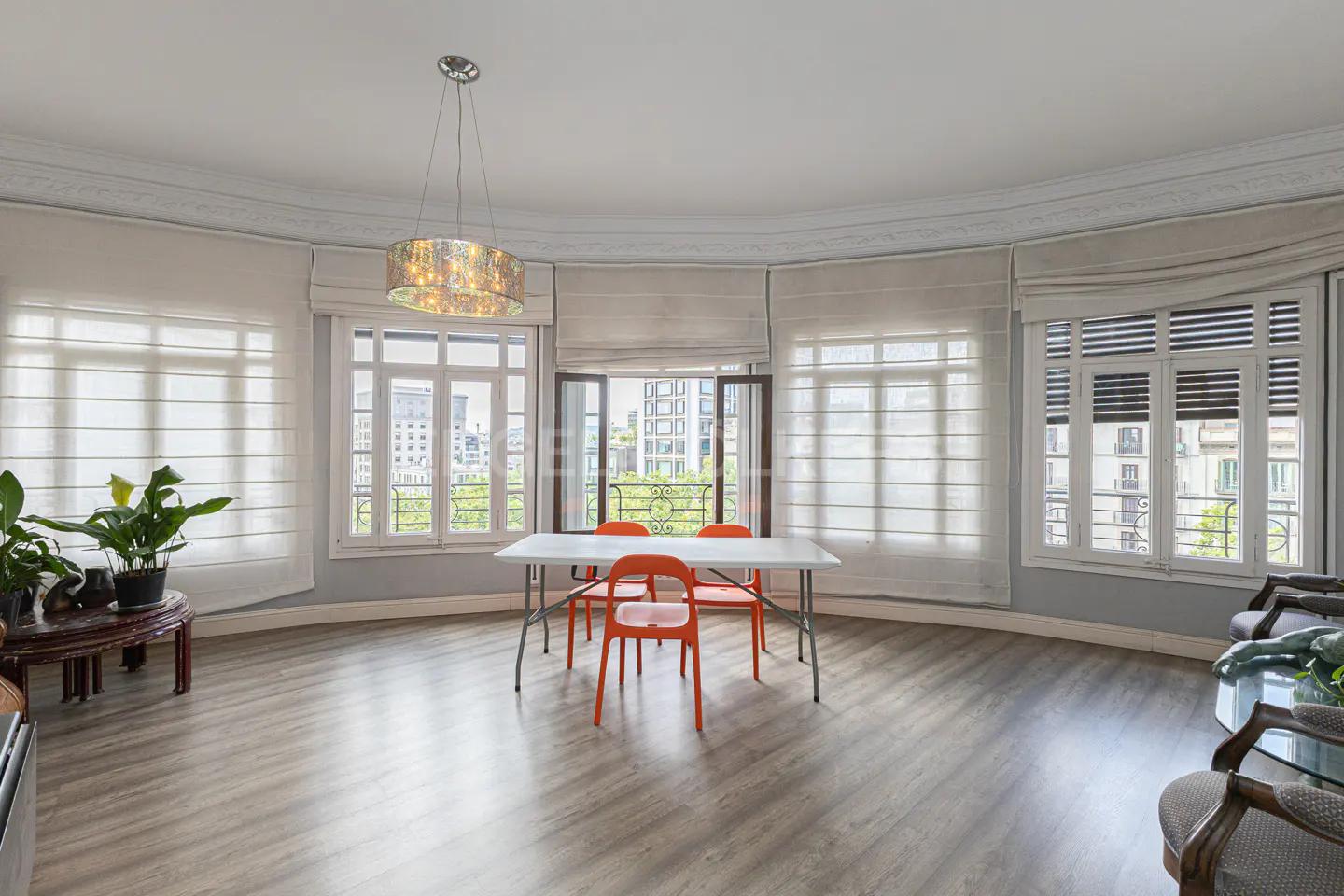 Bright dining room with wood floors, white walls, and large windows. A white table and orange chairs sit under a modern chandelier.