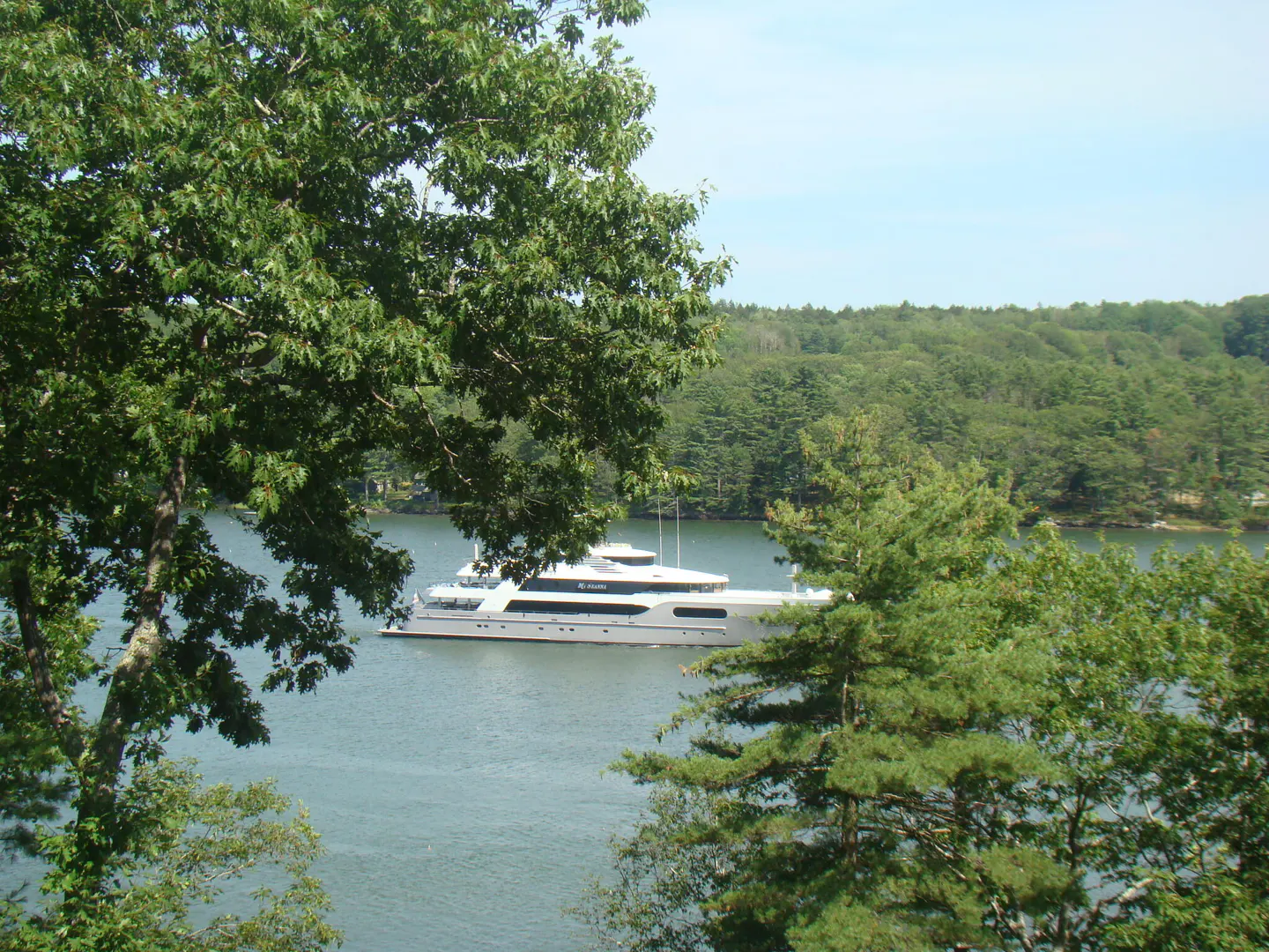 View of a large white yacht on a blue lake, framed by green trees and a forest in the background.