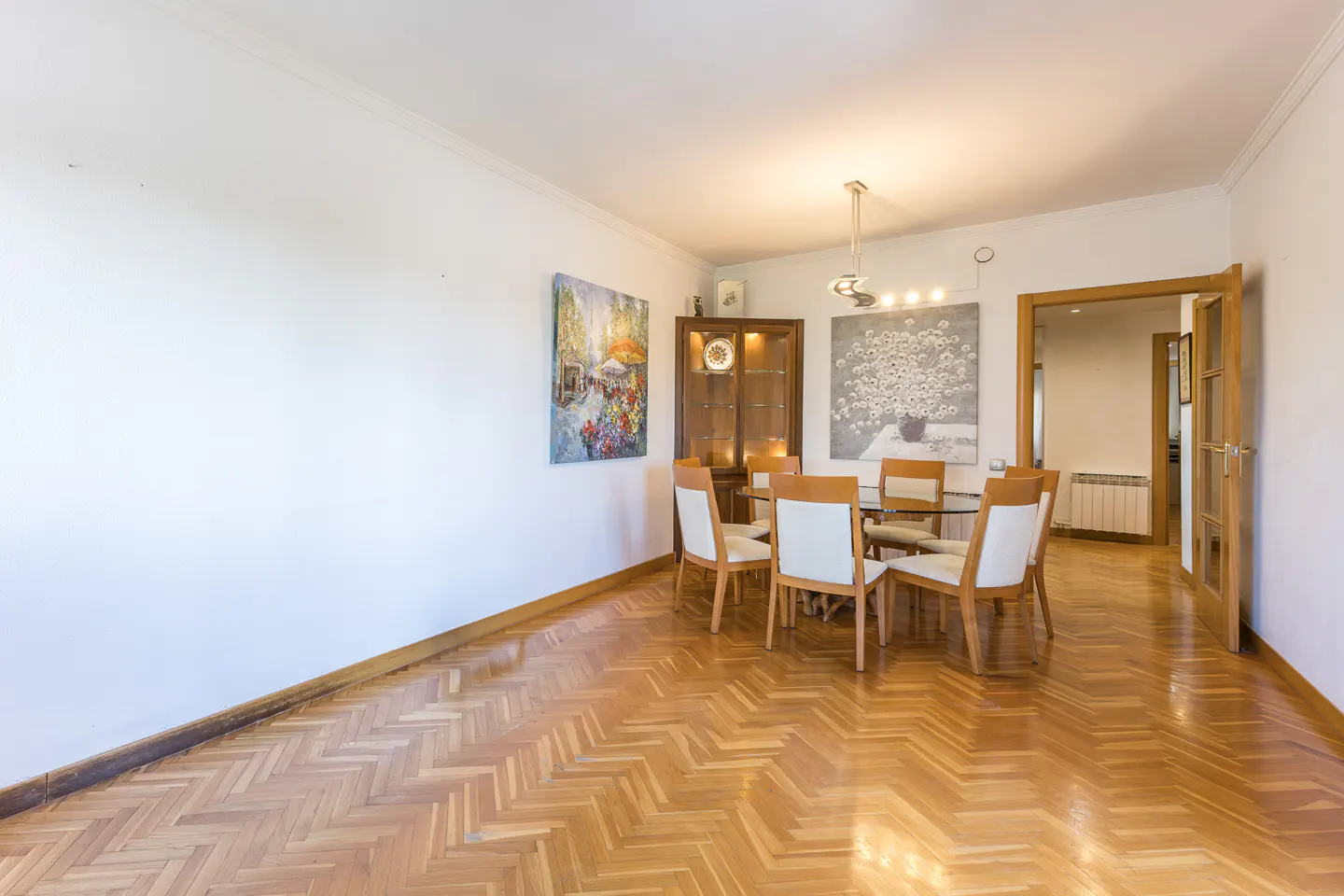 Bright dining room with parquet floors, white walls, and a glass table surrounded by six chairs. Artwork adorns the walls.