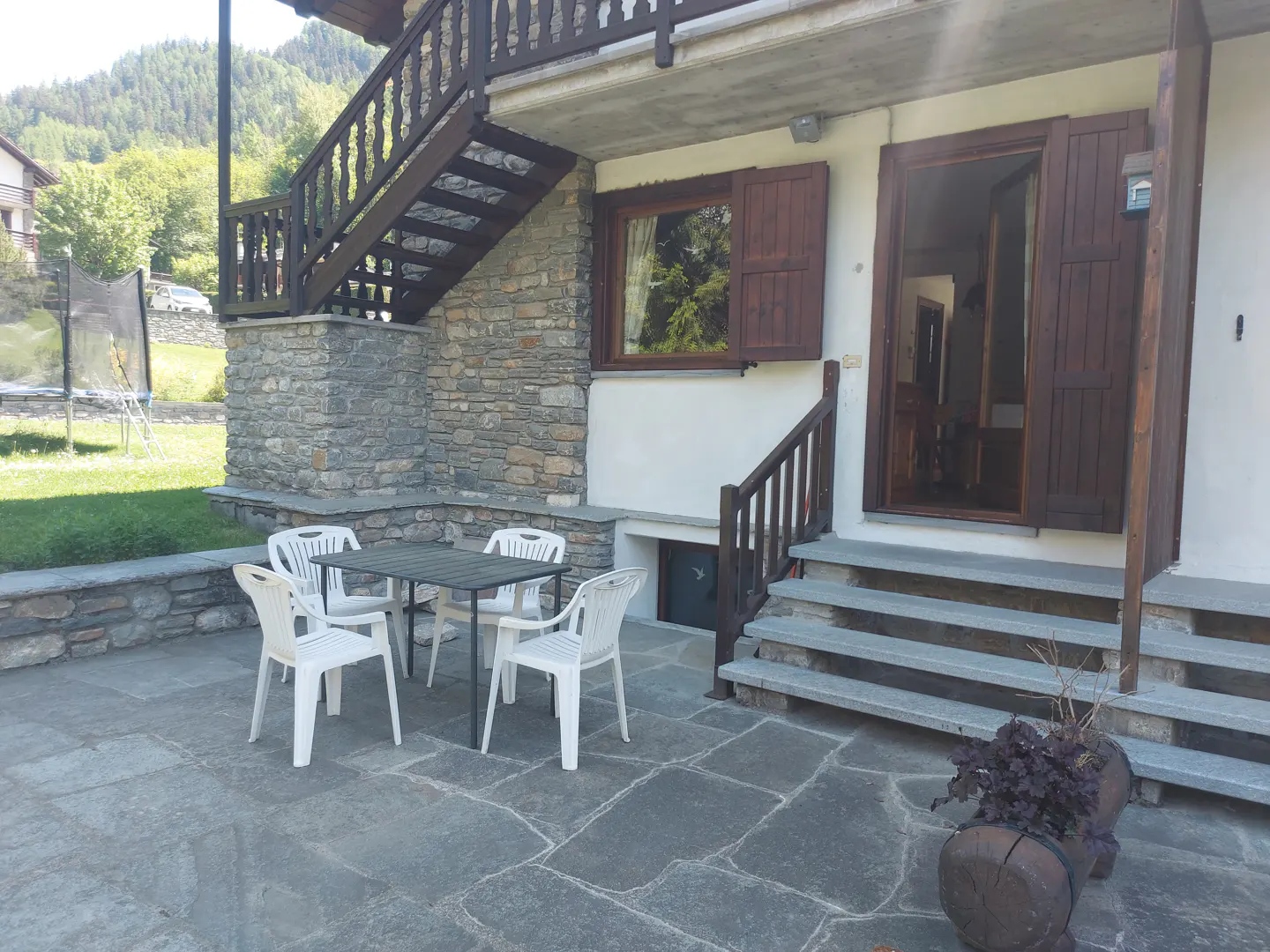 Stone patio with a table and four white chairs outside a house with stone walls, wooden shutters, and stairs.