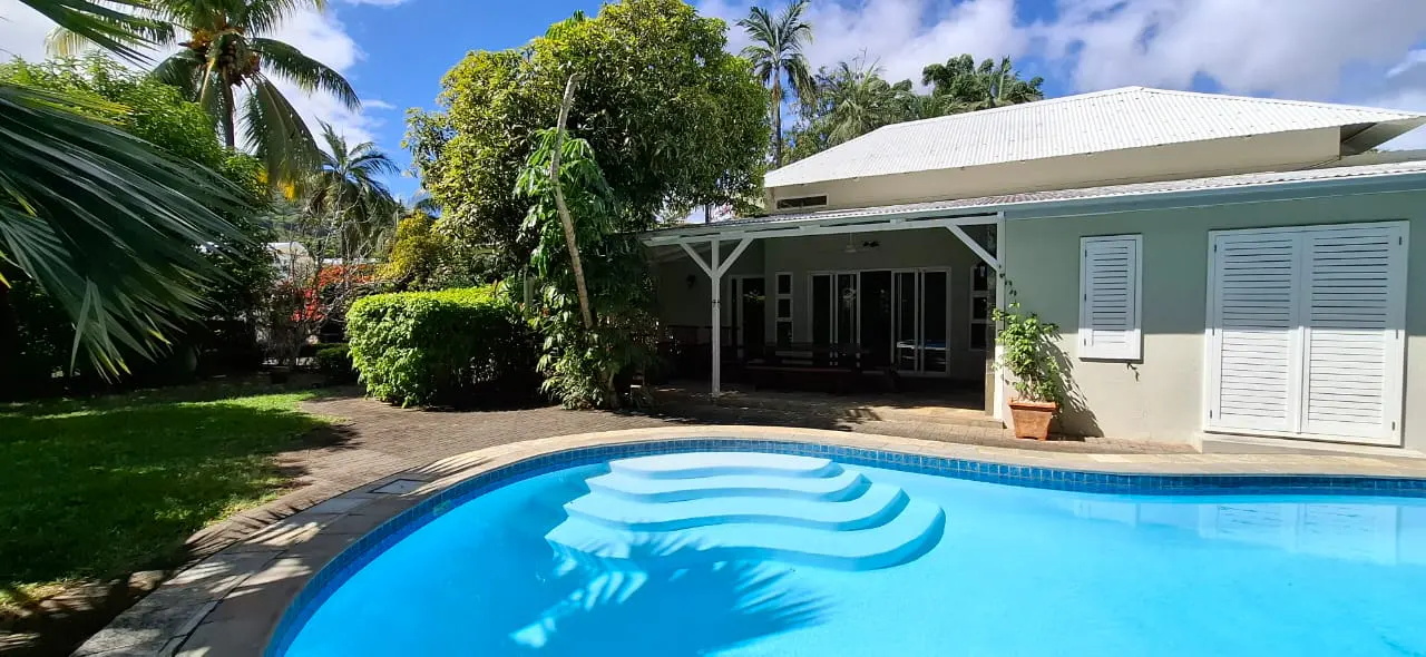 A light green house with a white roof and shutters sits beside a bright blue swimming pool with built-in steps.