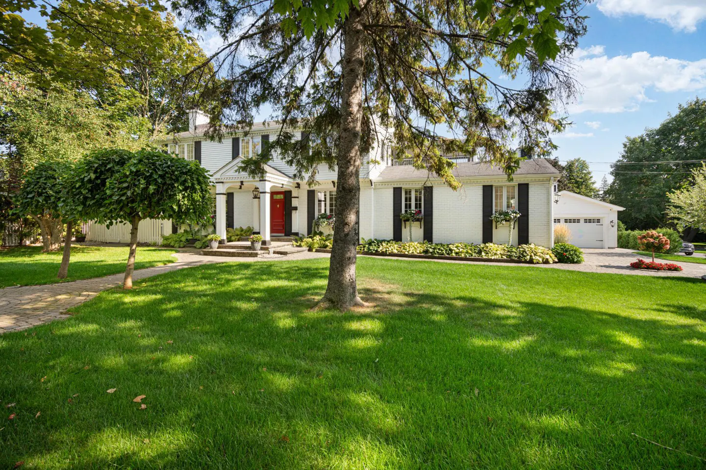 Two-story white house with black shutters and a red front door, surrounded by a green lawn and trees under a blue sky.