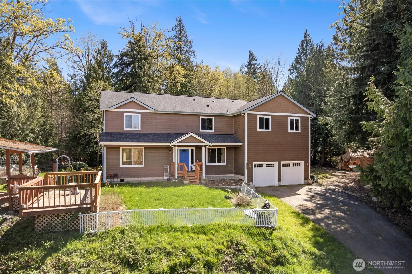 Two-story brown house with white trim, blue door, and two-car garage. Deck and green lawn in front, surrounded by trees.