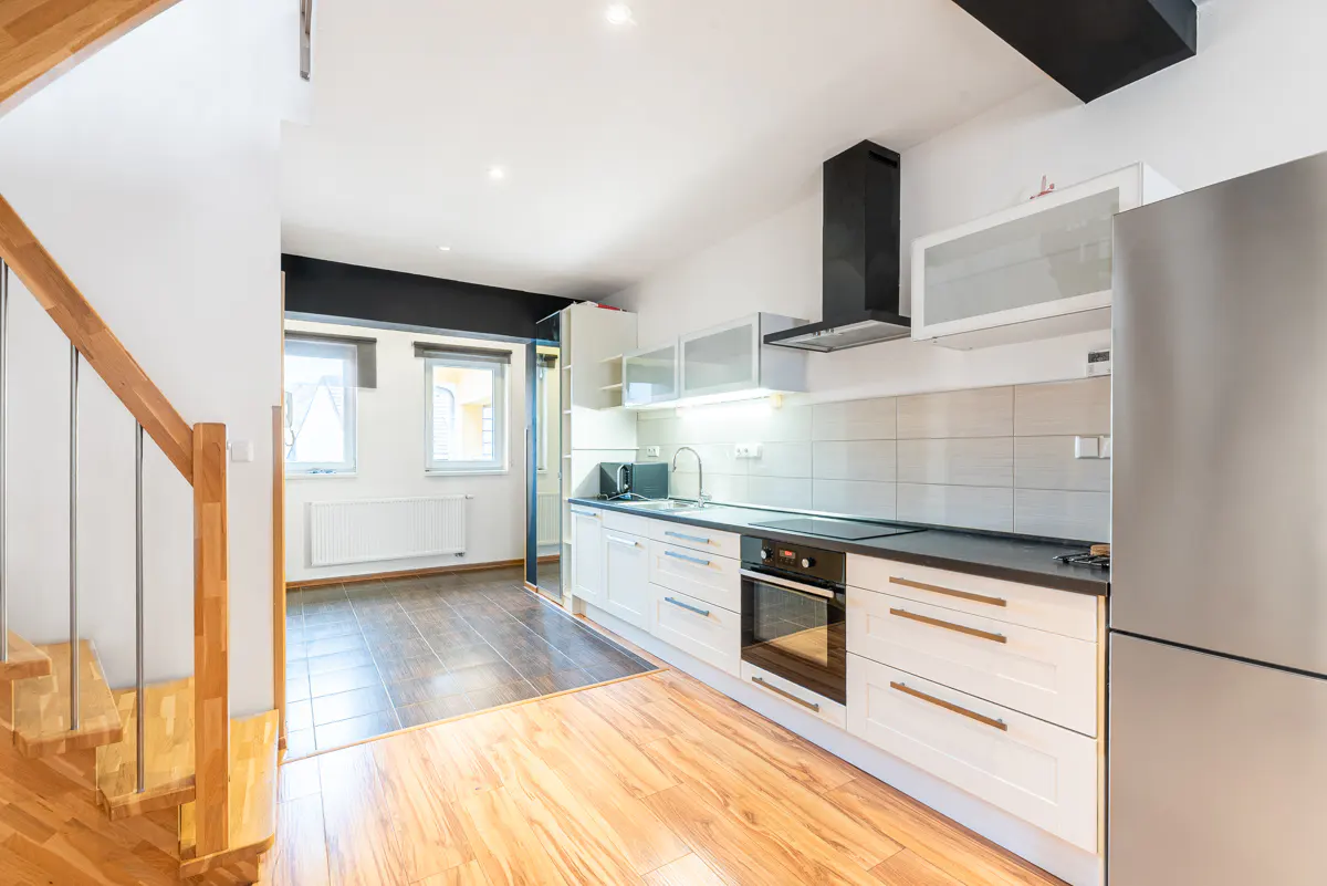 Bright kitchen with white cabinets, black countertops, and stainless steel fridge. Wood floors lead to a staircase on the left.