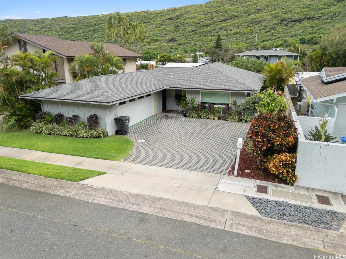 A single-story home with a gray roof and light gray siding, a patterned driveway, and a green, grassy hill in the background.