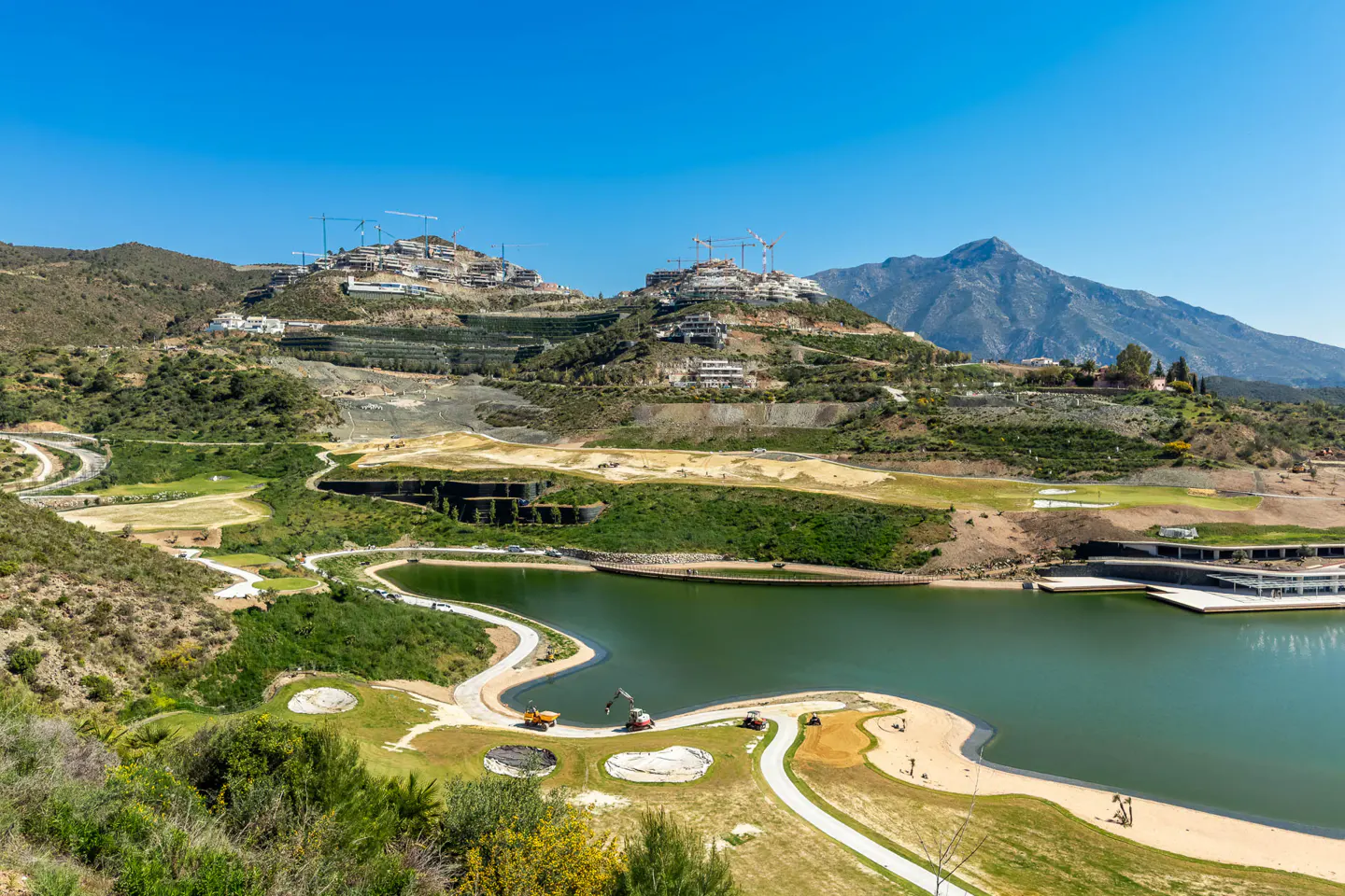 Scenic view of a lake, golf course, and hillside homes under construction on a sunny day.