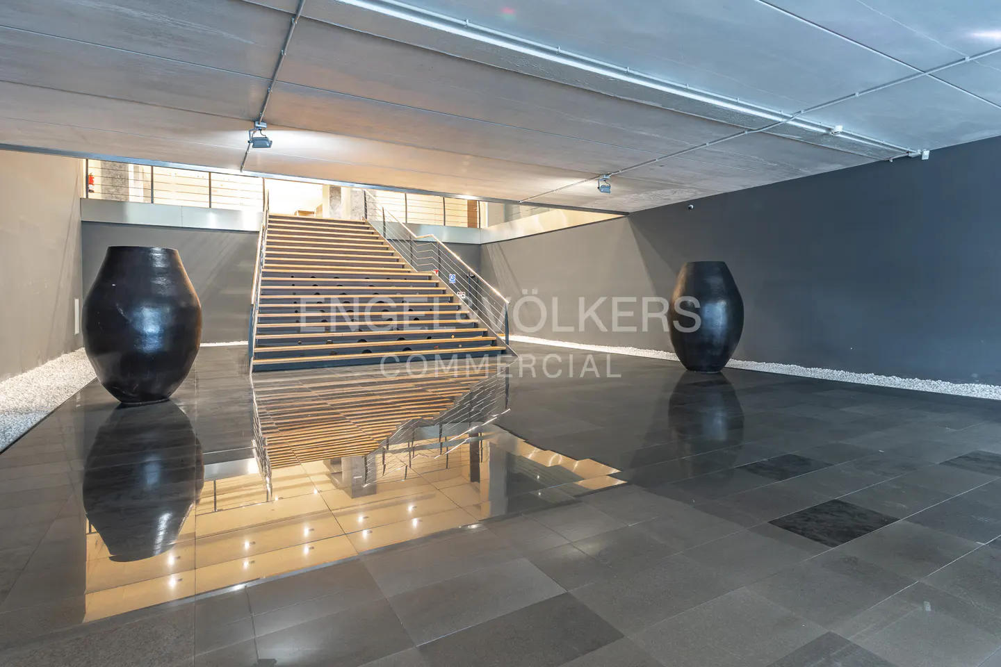 Modern lobby with black tile floor, stairs, and two large black vases. The ceiling is concrete with recessed lighting.