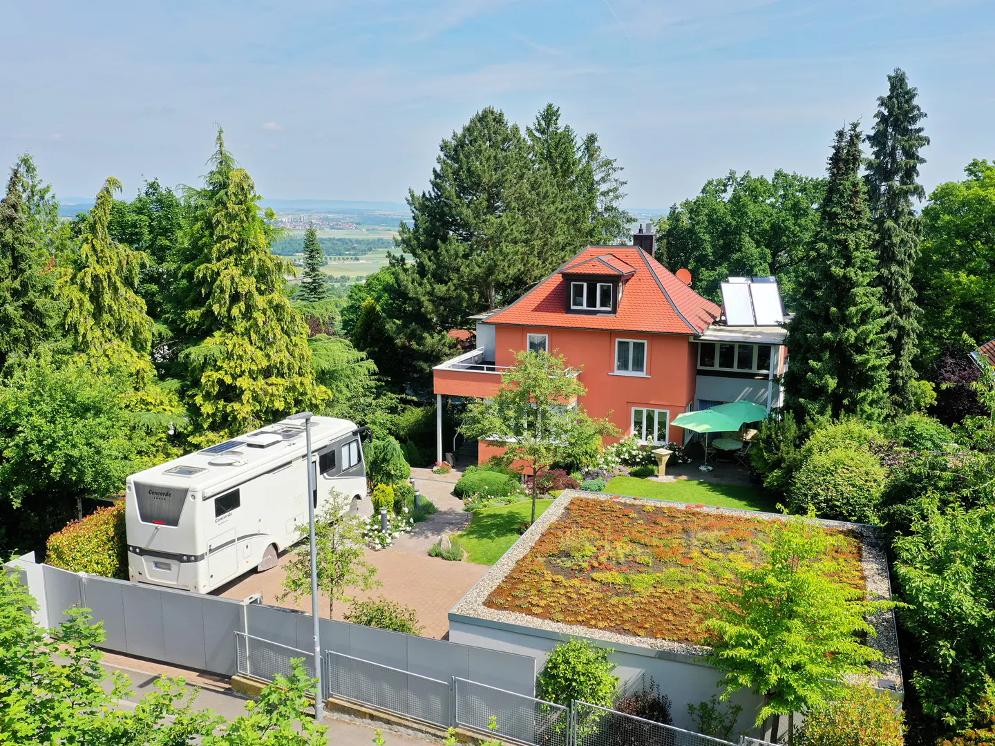 Aerial view of a two-story orange house with a red tile roof, surrounded by lush green trees and a white RV parked nearby.