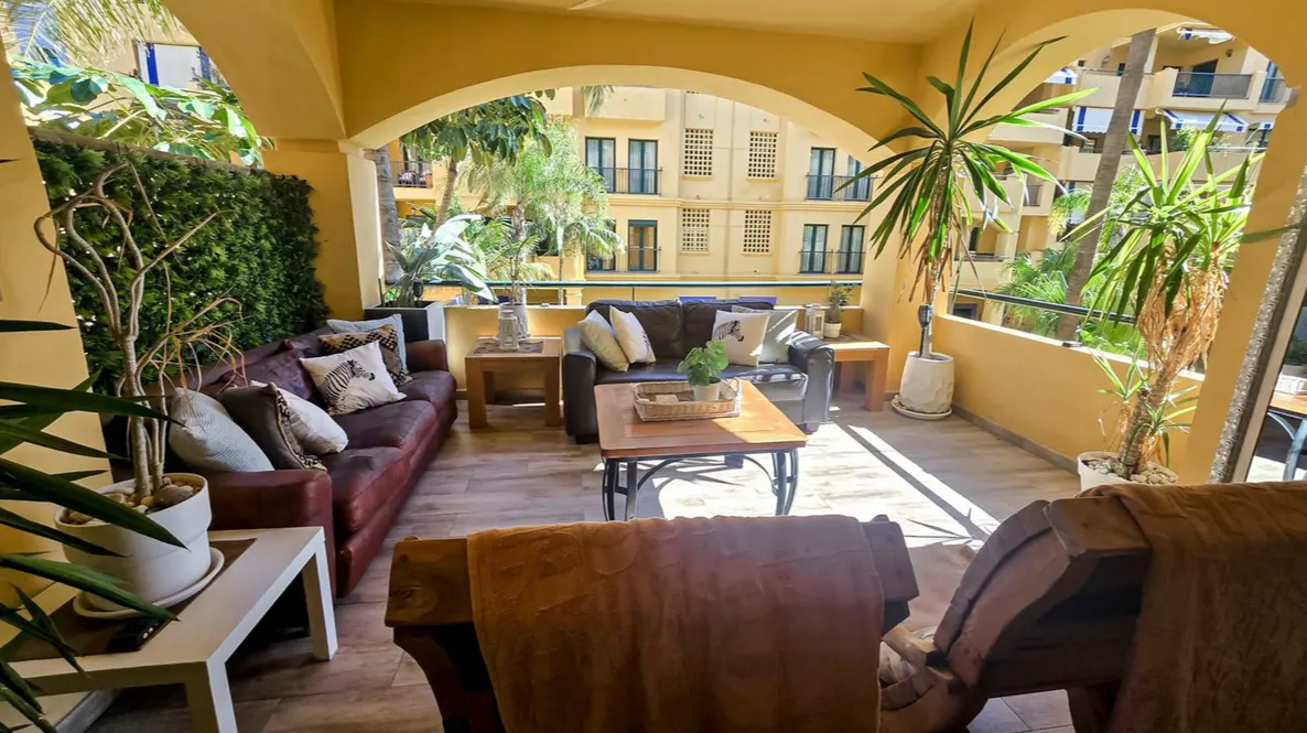 Covered balcony with brown sofas, wooden coffee table, potted plants, and a view of a yellow apartment building.