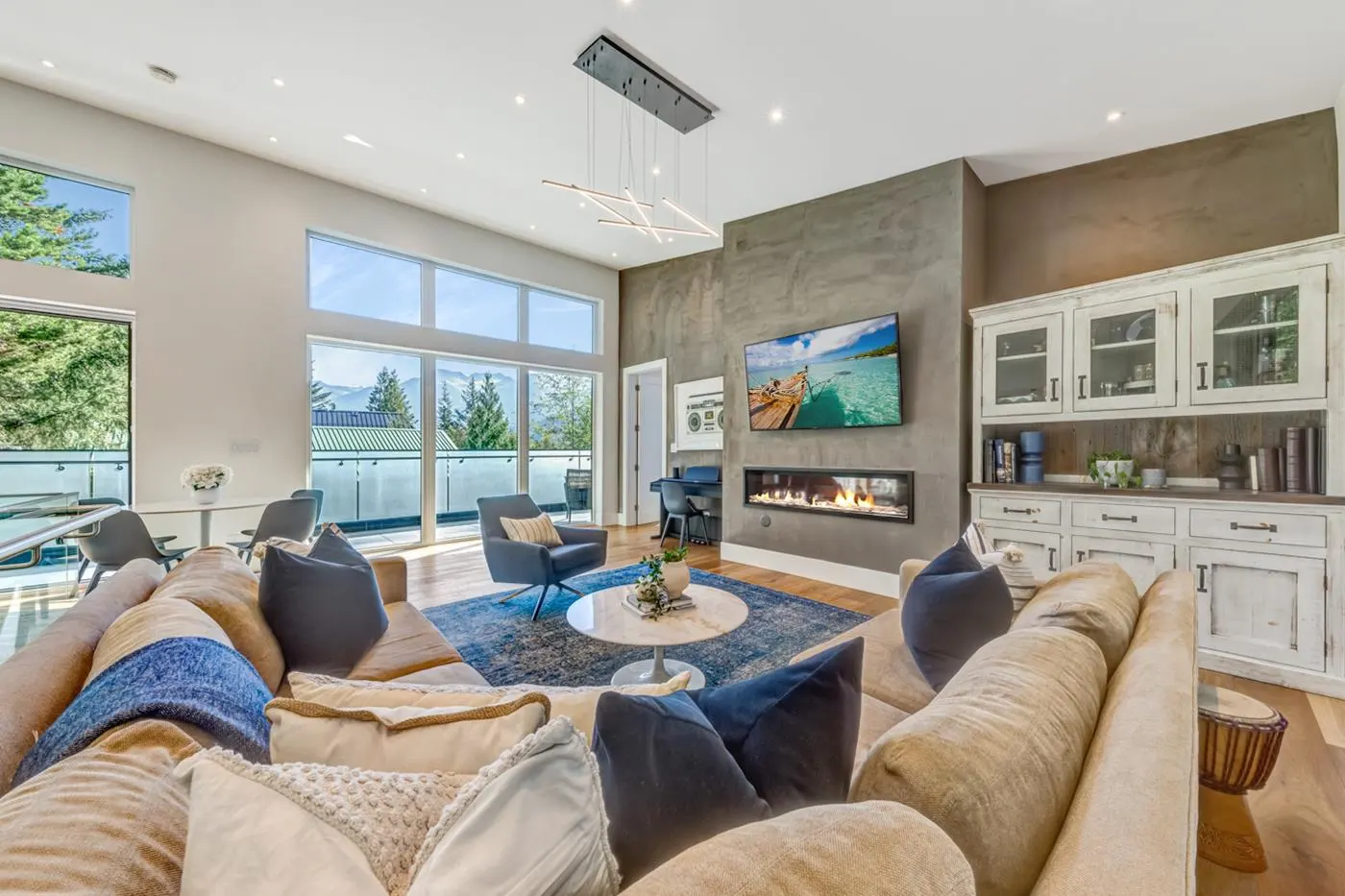 Bright living room with a large beige sectional sofa, blue rug, fireplace, TV, and floor-to-ceiling windows with mountain views.