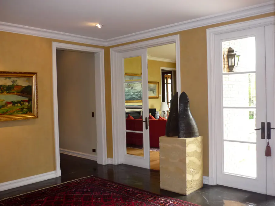 A foyer with gold walls, white trim, and dark floors. A red rug sits on the floor. A painting hangs on the wall. A sculpture sits on a pedestal.