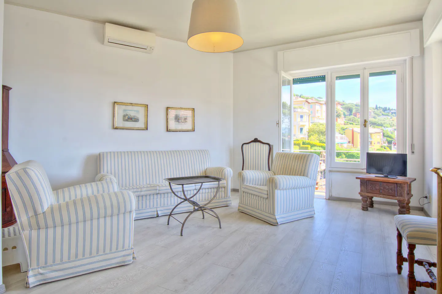 Bright living room with white walls, light wood floors, and striped furniture. A window shows a view of a town on a hill.