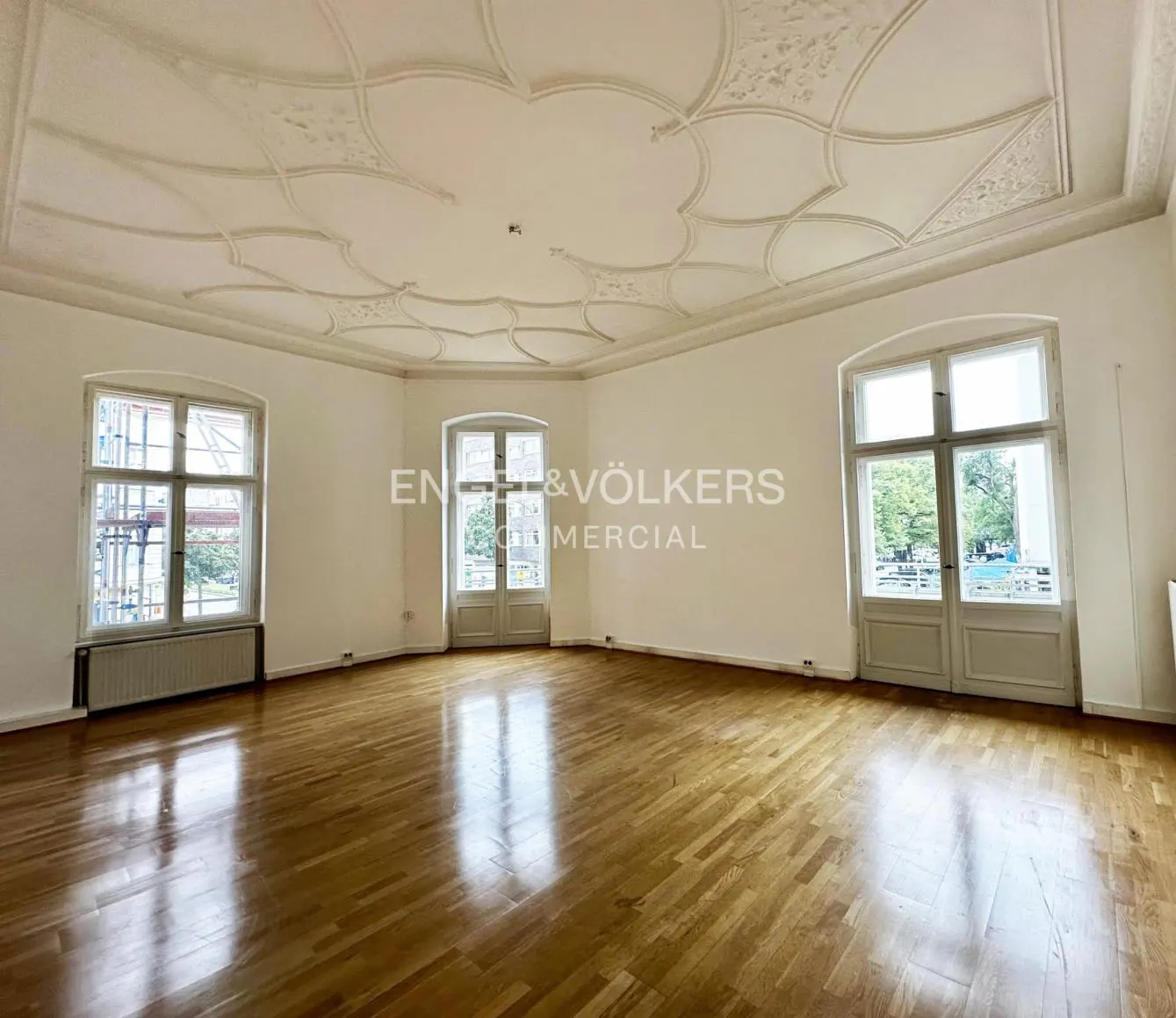 Empty room with hardwood floors, white walls, and decorative white ceiling. Three windows let in natural light.