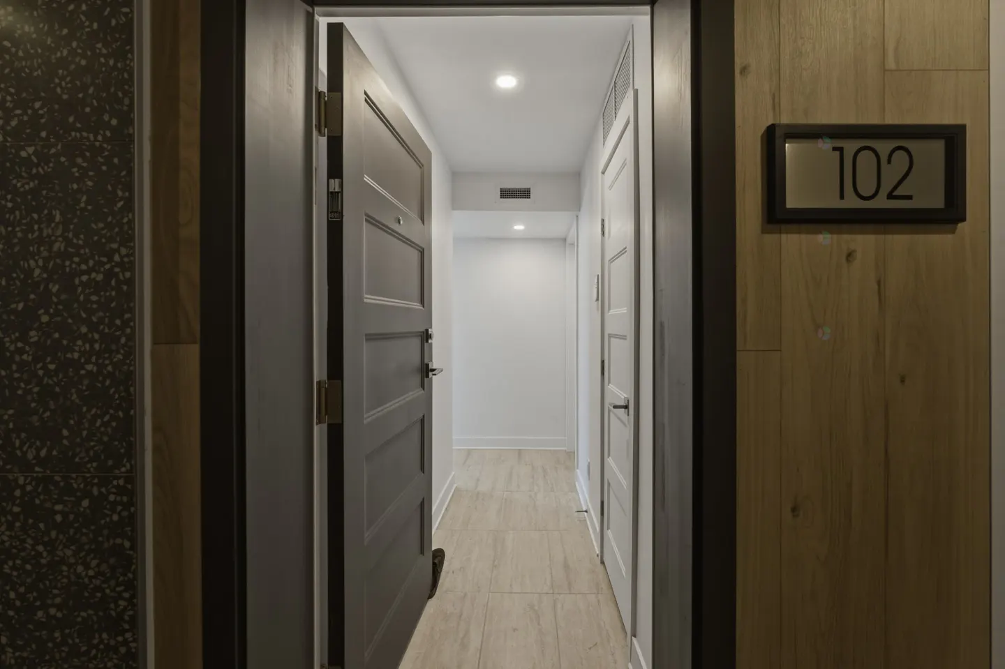 Hotel hallway with an open gray door to room 102. A white door is visible down the hall. The walls are wood and speckled black.