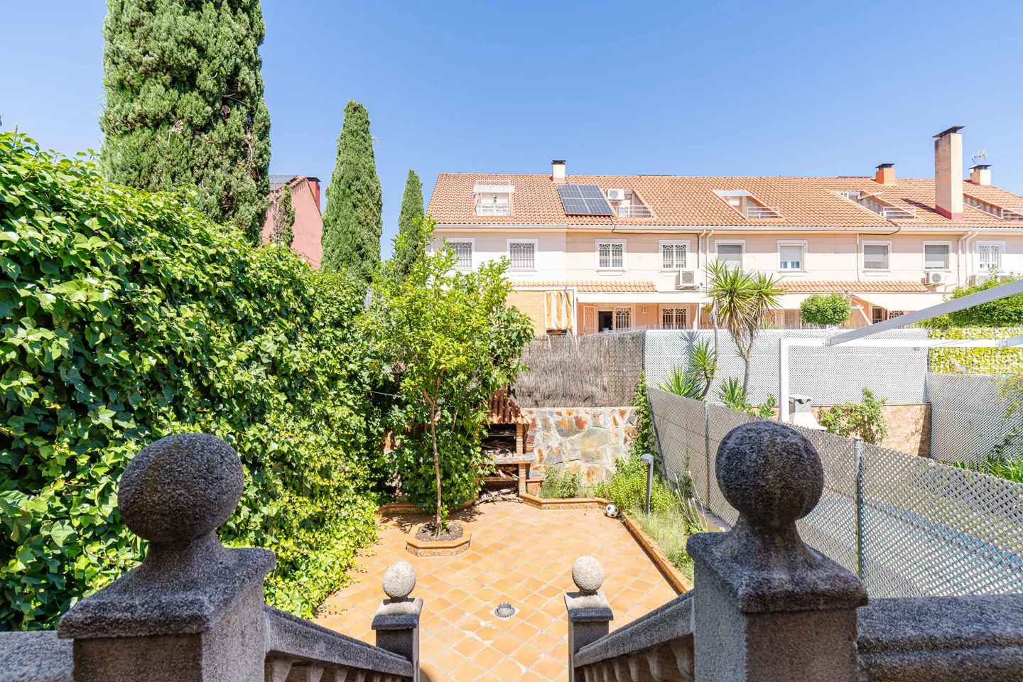 View from stone stairs overlooking a terracotta patio, a small tree, a brick barbecue, and a row of beige townhouses.