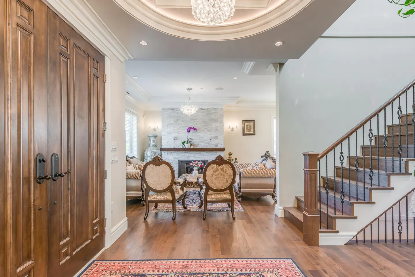 Grand foyer with dark wood double doors, hardwood floors, and staircase. Living room visible with fireplace and ornate furniture.