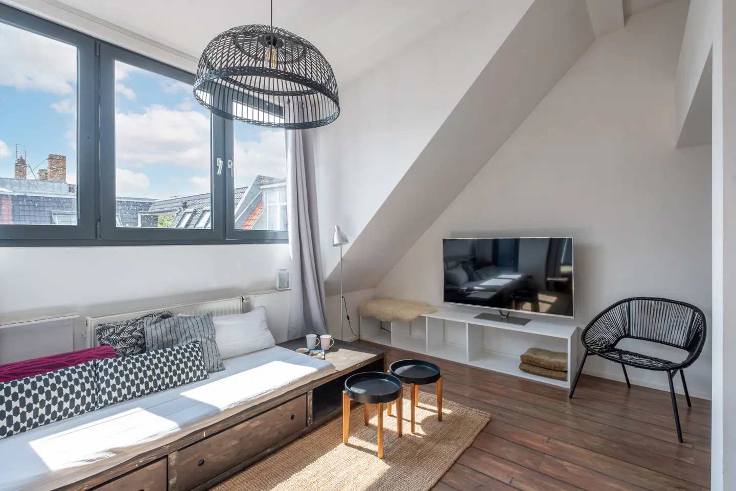 Attic living room with a daybed, pillows, and a black wicker pendant light. A TV sits on a white shelf under the sloped ceiling.