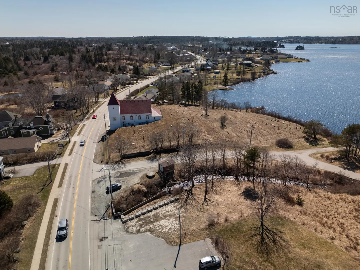 Aerial view of a white church with a red roof, a road with cars, and a lake in the background.