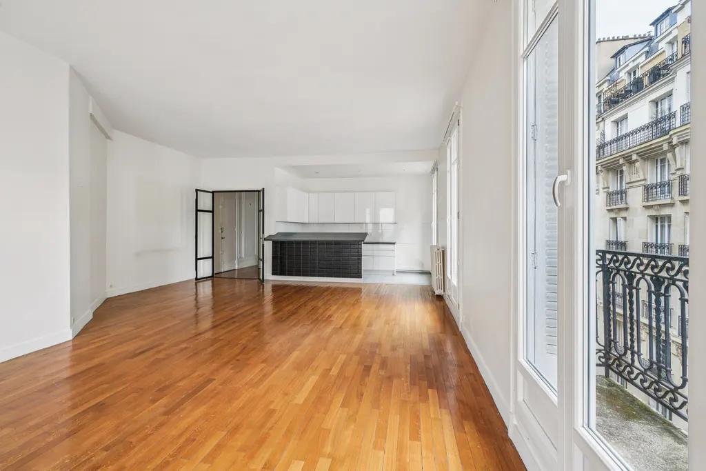 Bright, empty apartment with hardwood floors, white walls, and a kitchen with a black island. Balcony view of a Parisian building.