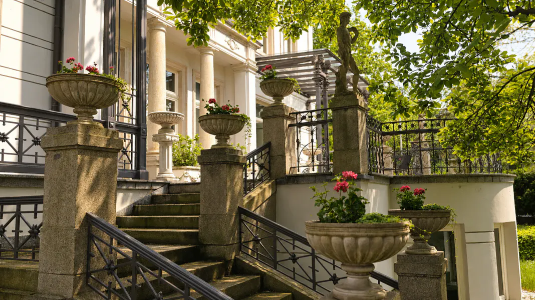 Stone steps lead to a white building with columns. Urns with red flowers line the stairs and a black metal railing. Green trees are in the background.