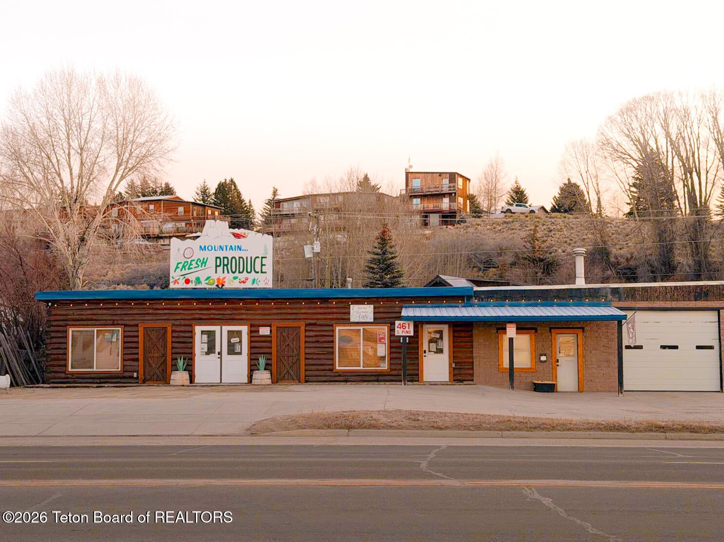 A brown log-style commercial building with a "Mountain Fresh Produce" sign, set against a hillside with houses at sunset. [AI-generated text]