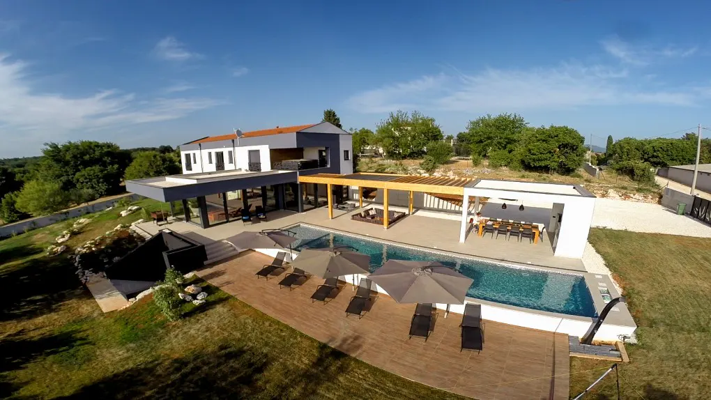 Aerial view of a modern two-story house with a pool, umbrellas, and lounge chairs on a sunny day.