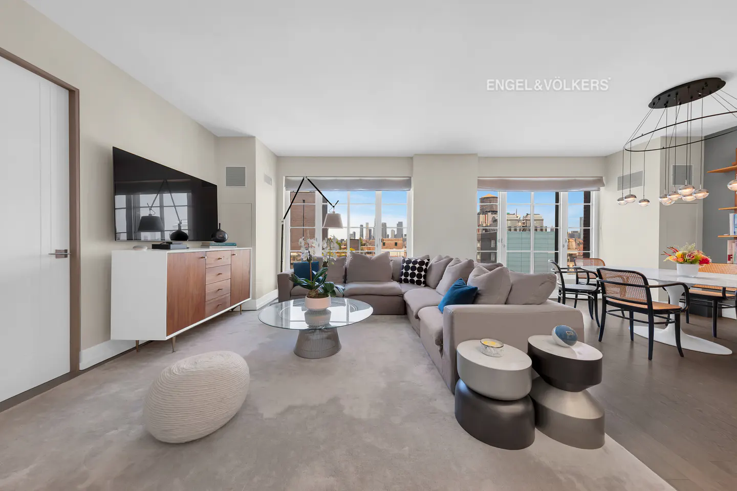 Living room with gray sectional sofa, glass coffee table, and city view through windows. Dining area with modern chandelier.