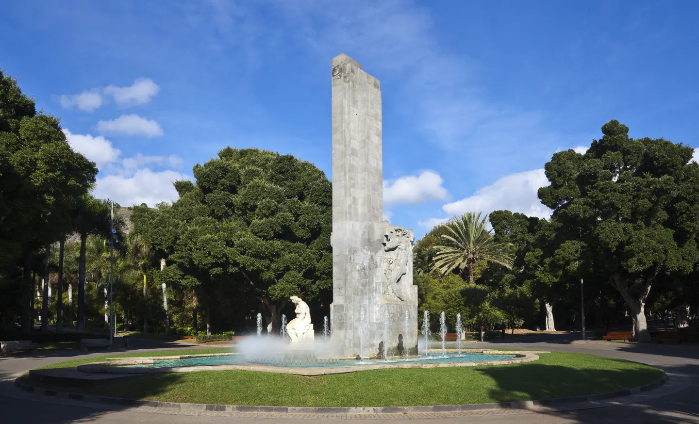 Park scene with tall monument, fountain, and lush trees under a blue sky with clouds.
